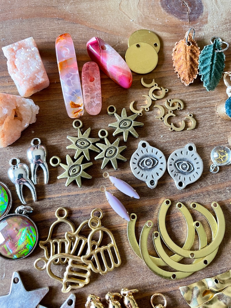 May include: A collection of jewelry-making supplies, including various charms, beads, and pendants. The assortment features gold-toned crescent moons, starbursts, and Hamsa hand charms. Also included are polished stones in shades of pink and orange, and leaf-shaped pendants.