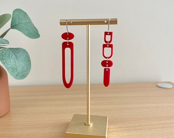 May include: A pair of red geometric earrings displayed on a gold-colored earring stand. One earring is a long, open rectangle, and the other is a series of connected shapes. The earrings are made of a solid, opaque material.