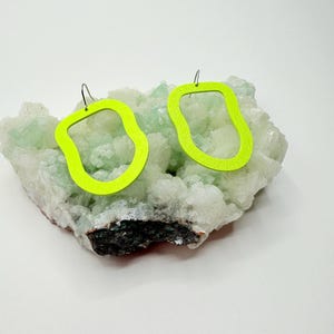 Puede incluir: Un par de pendientes de color verde neón con una forma orgánica única. Los pendientes están hechos de un material ligero y tienen un cierre de gancho plateado.