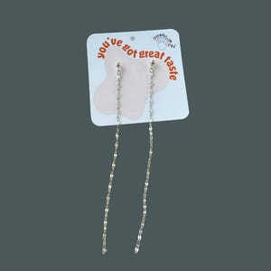 May include: Long, silver-coloured chain earrings with a slender, flowing design. The earrings are presented on a white card with the text "you've got great taste" in red and a small pineapple logo. The background is a muted grey.