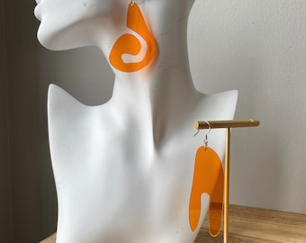 May include: A pair of orange abstract acrylic earrings. The earrings are shaped like a teardrop with a curved line inside. The earrings are hanging from a white mannequin head and a gold stand.