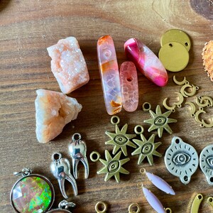 May include: A collection of jewelry-making supplies, including various gemstones, metal charms, and pendants. The gemstones are in shades of pink, orange, and clear. Charms include stars, moons, and teeth. The background is a wooden surface.