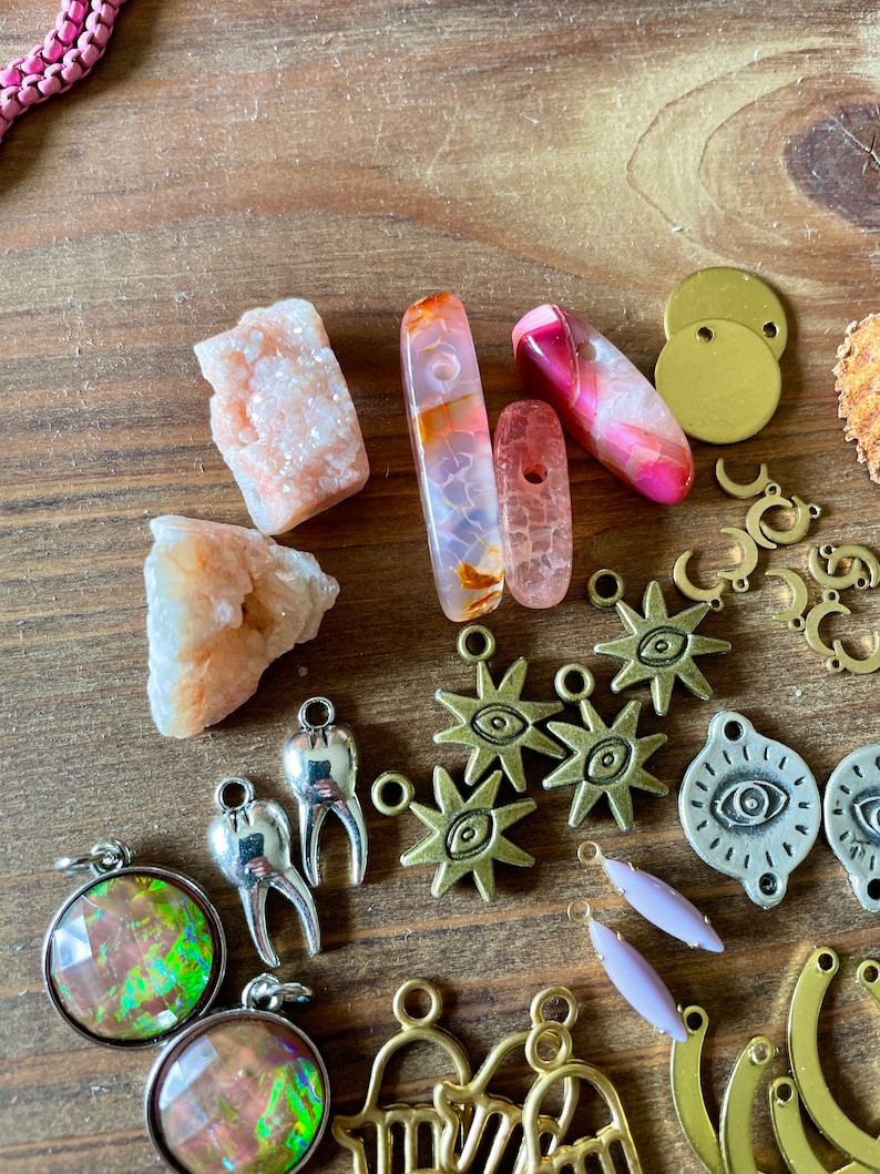 May include: A collection of jewelry-making supplies, including various gemstones, metal charms, and pendants. The gemstones are in shades of pink, orange, and clear. Charms include stars, moons, and teeth. The background is a wooden surface.