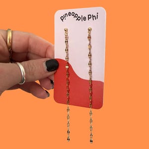 May include: Gold-colored chain earrings with a delicate design. The earrings are displayed on a white and red card with the text "pineapple phi" in black. The background is a solid orange color.