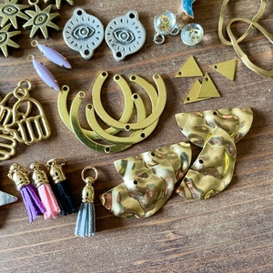 May include: An assortment of jewelry-making components, including gold-toned charms in various shapes: sunbursts, crescent moons, triangles, and hammered metal pieces. Also, there are silver-toned eye-shaped charms, lavender-colored teardrop beads, and colorful tassels.