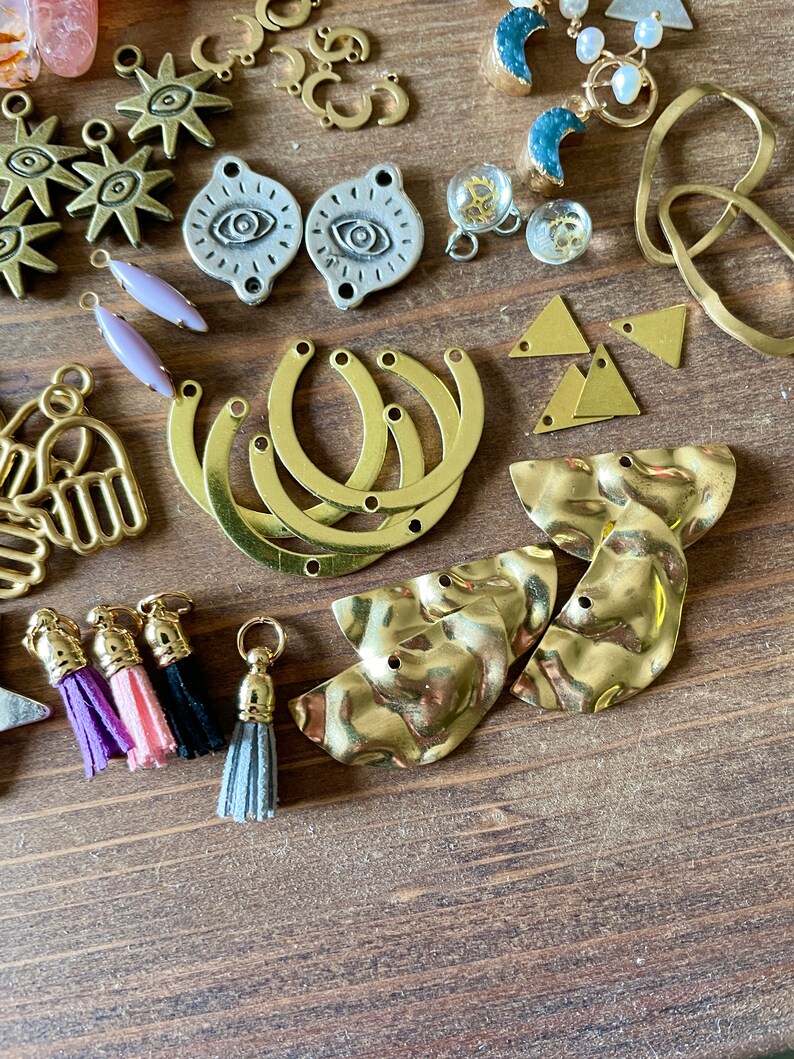 May include: An assortment of jewelry-making components, including gold-toned charms in various shapes: sunbursts, crescent moons, triangles, and hammered metal pieces. Also, there are silver-toned eye-shaped charms, lavender-colored teardrop beads, and colorful tassels.