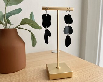 May include: Black geometric earrings with three different shapes on a gold stand. The earrings are made of black acrylic and have a modern, minimalist design.