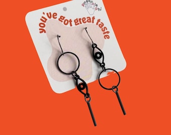 Puede incluir: Pendientes de metal negro con diseño circular y en forma de ojo, colgados de una tarjeta blanca con el texto rojo "you've got great taste". Los pendientes tienen un estilo minimalista y geométrico, sobre un fondo naranja.