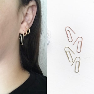 May include: A close-up of a person's ear with three gold hoop earrings. The earrings are different sizes and styles. The image also shows four gold paper clips on a white background.
