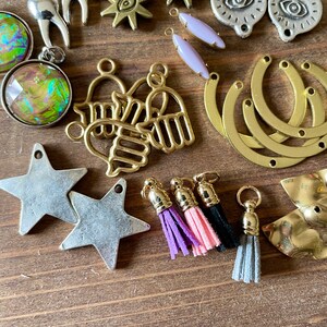 May include: A collection of jewelry-making components, including star-shaped charms, gold-toned hand charms, and eye-shaped pendants. Also shown are small tassels in purple, pink, black, and gray, along with other metal findings. The items are arranged on a wooden surface.