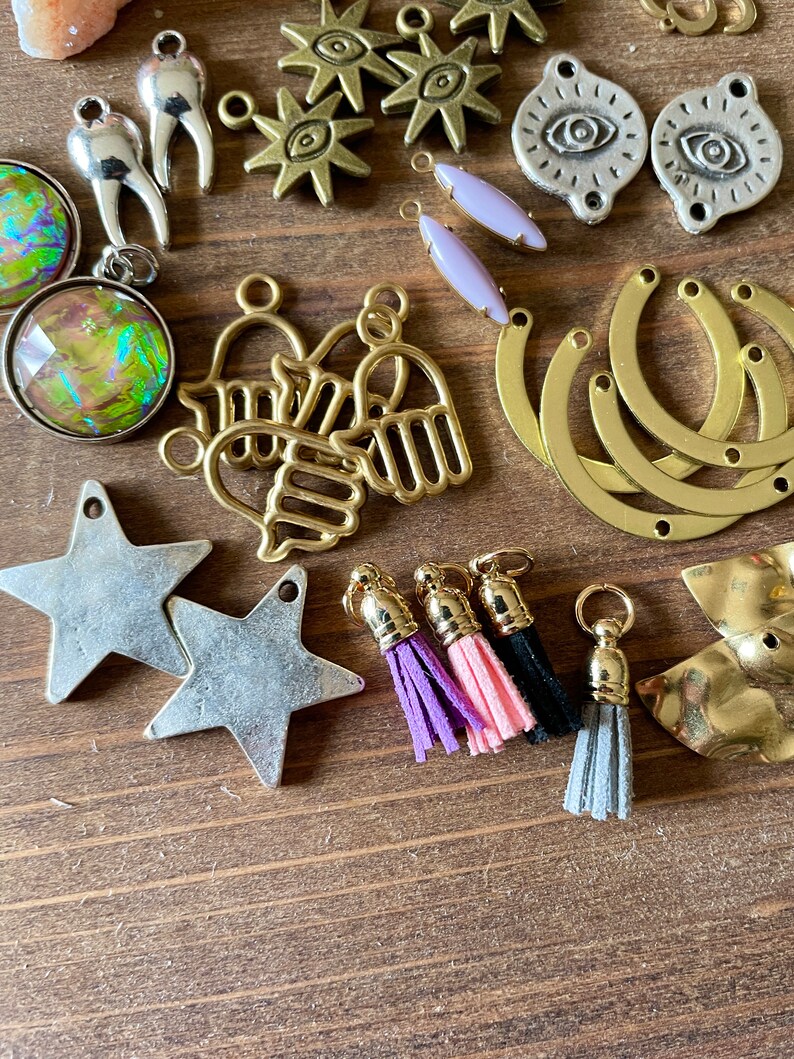 May include: A collection of jewelry-making components, including star-shaped charms, gold-toned hand charms, and eye-shaped pendants. Also shown are small tassels in purple, pink, black, and gray, along with other metal findings. The items are arranged on a wooden surface.