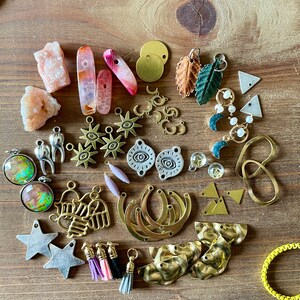 May include: A collection of jewelry-making supplies, including various charms, beads, and pendants. The assortment features gold-toned metal pieces, colorful gemstones, and small tassels. The items are arranged on a wooden surface, showcasing a variety of shapes and textures.