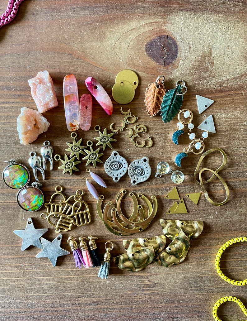 May include: A collection of jewelry-making supplies, including various charms, beads, and pendants. The assortment features gold-toned metal pieces, colorful gemstones, and small tassels. The items are arranged on a wooden surface, showcasing a variety of shapes and textures.