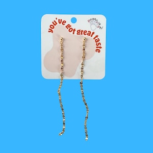May include: Gold-coloured dangle earrings with a delicate chain design. The earrings are attached to a white card with the text "you've got great taste" and a pineapple logo. The background is a solid blue.