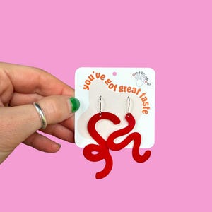 May include: Red, abstract-shaped earrings with silver hooks. The earrings are displayed on a white card with the text "you've got great taste" and a hand-drawn logo. The background is a solid pink.