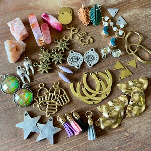 May include: A collection of jewelry-making supplies and components, including gemstones, metal charms, and tassels. The assortment features various shapes, colors, and textures, such as pink crystals, gold-toned pendants, and silver star charms. The items are arranged on a wooden surface.