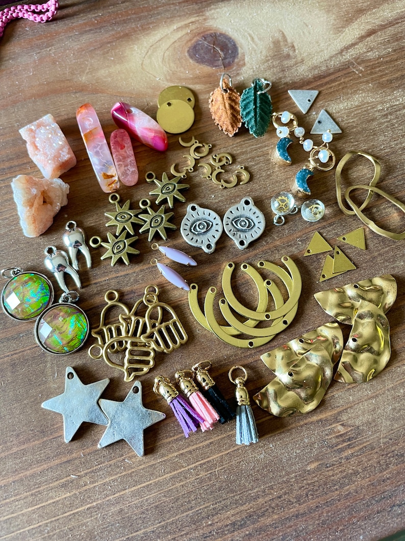 May include: A collection of jewelry-making supplies and components, including gemstones, metal charms, and tassels. The assortment features various shapes, colors, and textures, such as pink crystals, gold-toned pendants, and silver star charms. The items are arranged on a wooden surface.