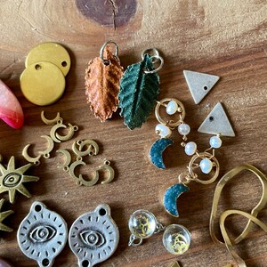 May include: An assortment of jewelry components and charms, including gold-colored round discs, crescent moons, leaf-shaped pendants, and eye-shaped charms. There are also earrings with crescent moon and pearl accents, and small glass globe earrings with gears inside.