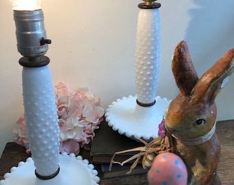 May include: Pair of white milk glass lamps with a beaded design and a white base. The lamps are lit and have a warm glow. The lamps are on a wooden surface with a brown bunny holding a pink Easter egg.