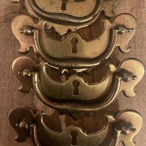 May include: Four antique brass drawer pulls with keyhole details are arranged vertically. The pulls have a curved handle design with decorative flourishes on each side. The brass has a slightly aged, tarnished appearance, suggesting vintage or reclaimed hardware.
