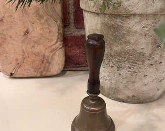 May include: A brass bell with a wooden handle. The bell is a light gold color and has a rounded shape. The handle is dark brown and has a simple design.