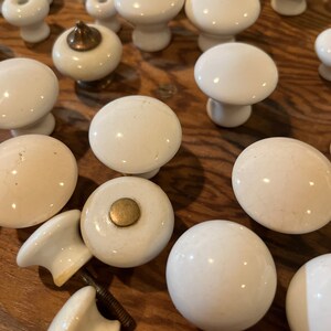 May include: A collection of white ceramic cabinet knobs with a variety of styles, including round, mushroom-shaped, and ball-shaped. Some knobs have a gold accent.