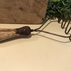May include: An antique potato masher with a wooden handle and a metal mashing head. The handle is painted white with visible wear and tear, and the metal shows signs of age and use. A rustic kitchen tool.