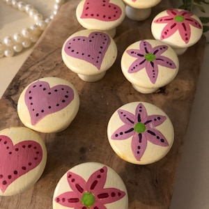 May include: A collection of hand-painted wooden cabinet knobs. The knobs feature heart and flower designs in shades of pink and purple, with green accents, set against a cream-colored background. They are arranged on a wooden surface.