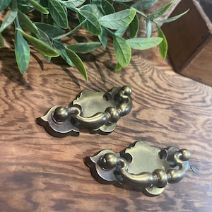 May include: Two antique brass drawer pulls with a decorative design. Each pull features a curved handle with ball accents and an ornate backplate. The hardware is set against a wooden surface, suggesting a vintage or rustic aesthetic.