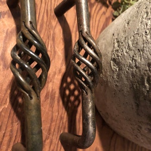 May include: Two antique bronze-colored metal handles. Each handle has a cage-like design in the center and a curved end. The handles are displayed on a wooden surface.