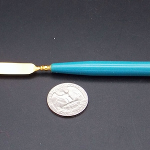 May include: A small, gold-colored butter knife with a teal and yellow handle. The knife is about the size of a US quarter.