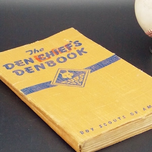 May include: A yellow book titled "The Den Chief's Denbook" with a blue and white Boy Scouts of America logo. The book is on a black surface with a baseball on a stand to the right.