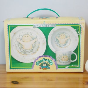 Peut inclure: Une boîte jaune avec une poignée verte contient un service de table Cabbage Patch Kids première édition. L'ensemble comprend deux bols blancs avec un motif Cabbage Patch Kid et une tasse blanche avec un motif Cabbage Patch Kid. La boîte porte les inscriptions "Royal Worcester" et "Première édition Cabbage Patch Kids".
