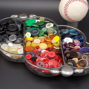 May include: A glass dish shaped like a clover leaf with three sections filled with colorful buttons. The sections contain black, white, and clear buttons, green, yellow, orange, and red buttons, and blue, purple, and brown buttons.