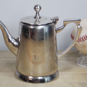 May include: A silver teapot with a shiny finish and a rounded body. The teapot has a lid with a knob on top and a handle on the side.