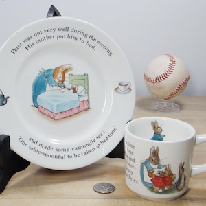 May include: A set of two white ceramic plates featuring illustrations from the children's book "Peter Rabbit". The larger plate has a quote from the book and a picture of Peter Rabbit in bed. The smaller plate has a picture of three rabbits.