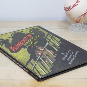 May include: A black hardcover book titled "Frankenstein Moved in on the Fourth Floor" by Elizabeth Levy and illustrated by Mordicai Gerstein. The cover features a green and yellow illustration of a monster with two children on a railing.