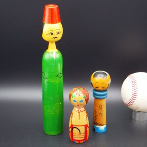 Puede incluir: Tres muñecas de madera con caras pintadas y ropa colorida, junto con una pelota de béisbol blanca con costuras rojas.