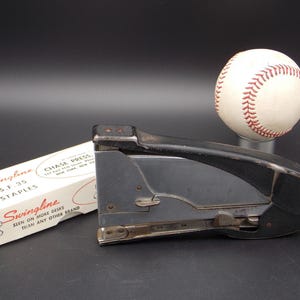 May include: A black Swingline stapler with a black handle and a silver metal base. The stapler is sitting on a white box with red text that reads "Swingline 5.F.35 Staples" and "Chase Press. 133 East 47th Street, New York, New York". The box also has the text "Swingline Seen on More Desks Than Any Other Brand". A white baseball with red stitching is sitting on a black surface behind the stapler.