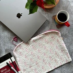 Op de afbeelding: Een zilveren laptop, een gewatteerde hoes met wit en roze patroon, een wijnjournaal, een koffiemok en een poinsettia-plant liggen op een grijze ondergrond. De laptop heeft een zwart Apple-logo. Het journaal heeft de tekst "Wine Journal Carnet Vins".