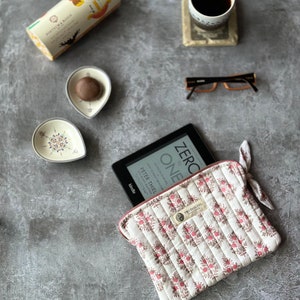 May include: A white quilted e-reader case with a pink floral pattern and a zipper closure. The case has a small label with the text "TRO PAISLETS" on it. The case is lying on a gray surface with an e-reader, a cup of coffee, a small plate with a chocolate candy, and a pair of glasses.