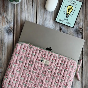 May include: A quilted laptop sleeve in a soft pink fabric featuring a repeating floral pattern of small red flowers and green leaves. The sleeve has vertical stitching, a zip closure with a fabric pull, and a small rectangular brand label.