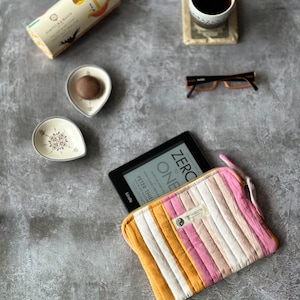 May include: A striped fabric e-reader case with a zipper closure. The case is yellow, pink, and white with a small brown label that says "Two Parleys Studio". The case is lying open on a grey surface with an e-reader inside. The e-reader screen shows the title "Zero to One" by Peter Thiel.