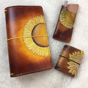May include: Three leather journals with sunflower designs. The largest journal is brown with a yellow sunflower design. The medium journal is brown with a yellow sunflower design. The smallest journal is brown with a yellow sunflower design.