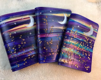 May include: Three purple leather journals with a moon and stars design. The journals have a watercolor effect with gold accents and a silver moon.