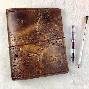 May include: A brown leather journal with a gold and brown embossed design of gears and clocks. The journal has red stitching and is closed. A clear pen and a white pencil are lying next to the journal.