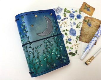 May include: A blue leather journal with a crescent moon and stars design. The cover is embossed with a floral pattern and has a black elastic closure.