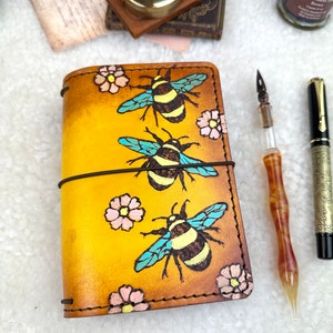 May include: A yellow leather journal with a floral and bee design. The journal has a brown leather strap and stitching. The bees are painted in blue, yellow, and black. The flowers are painted in pink and yellow.