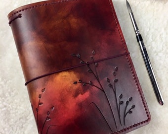 May include: A burgundy leather journal with a watercolor-like design and a floral pattern embossed on the cover. The journal is closed and lying on a white fuzzy surface.