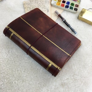 May include: A brown leather journal with gold accents and a burgundy elastic closure. The journal is closed and lying on a white surface.